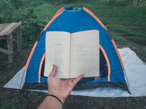 A person holds an open book in the foreground, with text visible on the pages. In the background, a blue and orange tent is set up on a white tarp on grassy ground. There's a wooden bench on the left side and lush greenery surrounding the area, giving a sense of being in a natural setting.