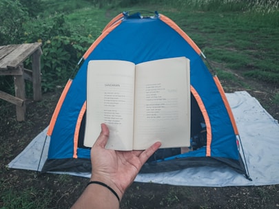 A person holds an open book in the foreground, with text visible on the pages. In the background, a blue and orange tent is set up on a white tarp on grassy ground. There's a wooden bench on the left side and lush greenery surrounding the area, giving a sense of being in a natural setting.