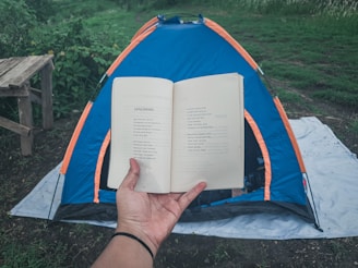 A person holds an open book in the foreground, with text visible on the pages. In the background, a blue and orange tent is set up on a white tarp on grassy ground. There's a wooden bench on the left side and lush greenery surrounding the area, giving a sense of being in a natural setting.