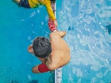 A person in a yellow shirt is assisting another person in red shorts who is swimming. The scene is set in a swimming pool with clear blue water. The perspective provides a bird's eye view focusing on the two individuals connected by a swimming lane divider.
