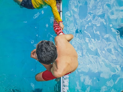 A person in a yellow shirt is assisting another person in red shorts who is swimming. The scene is set in a swimming pool with clear blue water. The perspective provides a bird's eye view focusing on the two individuals connected by a swimming lane divider.