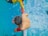 A person in a yellow shirt is assisting another person in red shorts who is swimming. The scene is set in a swimming pool with clear blue water. The perspective provides a bird's eye view focusing on the two individuals connected by a swimming lane divider.