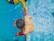 A person in a yellow shirt is assisting another person in red shorts who is swimming. The scene is set in a swimming pool with clear blue water. The perspective provides a bird's eye view focusing on the two individuals connected by a swimming lane divider.