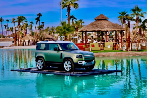 A green SUV is placed on a platform surrounded by a turquoise artificial lagoon, with palm trees and beach-style huts in the background under a clear blue sky.