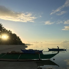 A serene beach scene at sunrise with two traditional boats on calm water, surrounded by lush palm trees. The sun is partially obscured by the trees, casting a warm glow across the scene. The horizon is visible with a clear sky dotted with scattered clouds.
