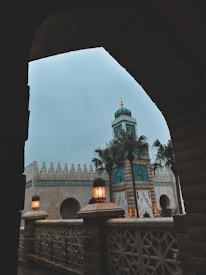 An architectural scene depicting a building with intricate details, featuring arched windows and a prominent tower. The tower is accented with turquoise and gold, complemented by palm trees nearby. Soft, warm lighting from lamps illuminates the foreground, creating a cozy atmosphere.