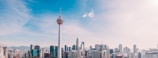 A city skyline featuring a prominent communication tower in the foreground, surrounded by modern skyscrapers under a clear blue sky.