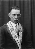 A black and white portrait of an older man with a serious expression. He has a short, neatly styled haircut and a thick mustache. He is wearing a suit with a decorated sash that features various symbols and text, including 'Past Chief Ruler' and other emblems.