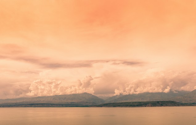 A serene landscape with a calm body of water in the foreground, expansive mountains in the middle ground, and a sky filled with soft, orange-hued clouds. The image exudes a peaceful, tranquil atmosphere with a wide, open view that suggests vastness and contemplation.