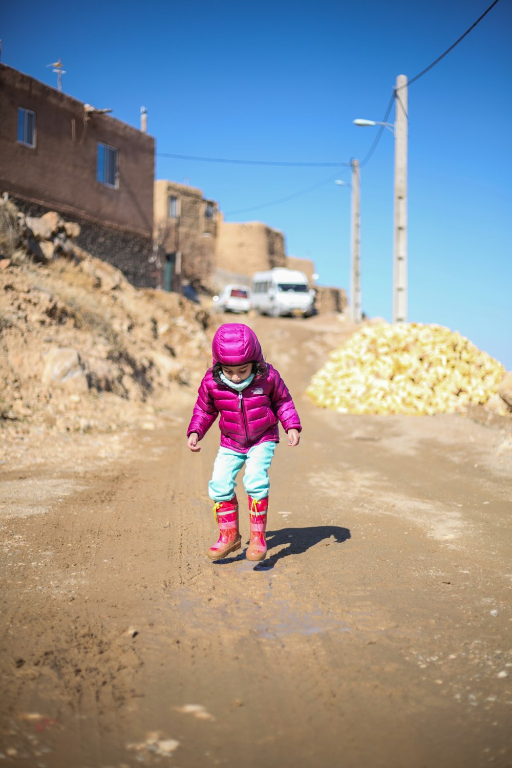 girl in pink jacket and blue pants running on brown dirt road during daytime