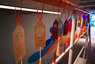 Several decorative tags hanging on a rail, each featuring intricate patterns and text in both Chinese and English. The tags are suspended by red strings and are adorned with tassels, creating a festive and cultural ambiance.