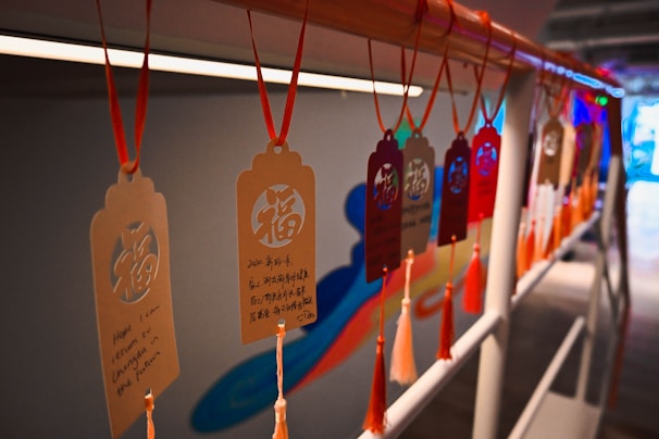 Several decorative tags hanging on a rail, each featuring intricate patterns and text in both Chinese and English. The tags are suspended by red strings and are adorned with tassels, creating a festive and cultural ambiance.