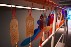 Several decorative tags hanging on a rail, each featuring intricate patterns and text in both Chinese and English. The tags are suspended by red strings and are adorned with tassels, creating a festive and cultural ambiance.