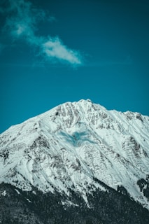 A majestic snow-covered mountain peak rises against a clear blue sky. Sparse clouds pass overhead, and a dense forest grows at the mountain's base.