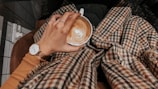 A hand adorned with modern aesthetic rings holding a cup of coffee, surrounded by a warm setting.