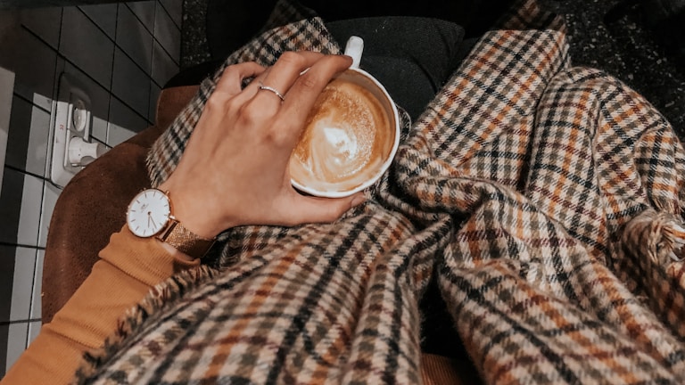 A hand adorned with modern aesthetic rings holding a cup of coffee, surrounded by a warm setting.