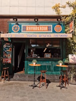 A small café with a vibrant, turquoise-painted wooden exterior and a vintage aesthetic. The signage features bold, colorful designs in orange and blue. Two small round tables with potted plants are positioned outside, each accompanied by wooden chairs. The setting feels inviting and warm, enriched by sunlight.