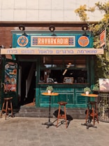 A small caf&eacute; with a vibrant, turquoise-painted wooden exterior and a vintage aesthetic. The signage features bold, colorful designs in orange and blue. Two small round tables with potted plants are positioned outside, each accompanied by wooden chairs. The setting feels inviting and warm, enriched by sunlight.