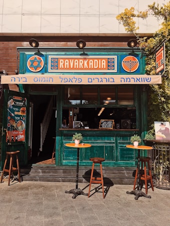 A small caf&eacute; with a vibrant, turquoise-painted wooden exterior and a vintage aesthetic. The signage features bold, colorful designs in orange and blue. Two small round tables with potted plants are positioned outside, each accompanied by wooden chairs. The setting feels inviting and warm, enriched by sunlight.