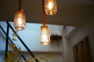 Three hanging light fixtures made from glass jars are suspended by ropes in a softly lit interior space. The light emitted casts a warm glow, complemented by yellow flowers in the foreground. An industrial staircase in the background enhances the modern yet rustic design.