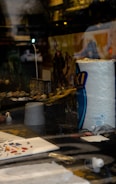 A display window features a variety of hobby items. There's a large roll of paper towels prominently visible. Posters or box art depict action scenes with fantastical characters in the background. Small, detailed figures or models are arranged, suggesting a focus on model painting or tabletop gaming. Paint palettes and brushes are scattered around.