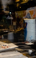 A display window features a variety of hobby items. There's a large roll of paper towels prominently visible. Posters or box art depict action scenes with fantastical characters in the background. Small, detailed figures or models are arranged, suggesting a focus on model painting or tabletop gaming. Paint palettes and brushes are scattered around.