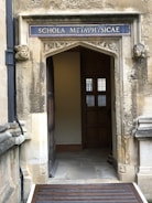 An ornate stone doorway with a sign reading 'SCHOLA METAPHYSICAE' in uppercase letters above. The doorway is framed by carved stone faces on each side. The interior visible through the doorway features wooden panels and a window with small panes. The stonework is weathered, giving a historical appearance.