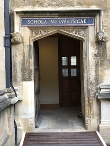 An ornate stone doorway with a sign reading 'SCHOLA METAPHYSICAE' in uppercase letters above. The doorway is framed by carved stone faces on each side. The interior visible through the doorway features wooden panels and a window with small panes. The stonework is weathered, giving a historical appearance.