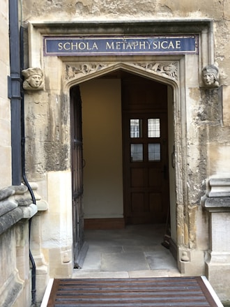 An ornate stone doorway with a sign reading 'SCHOLA METAPHYSICAE' in uppercase letters above. The doorway is framed by carved stone faces on each side. The interior visible through the doorway features wooden panels and a window with small panes. The stonework is weathered, giving a historical appearance.