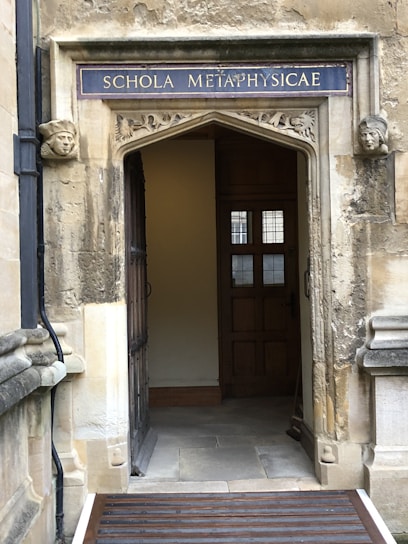 An ornate stone doorway with a sign reading 'SCHOLA METAPHYSICAE' in uppercase letters above. The doorway is framed by carved stone faces on each side. The interior visible through the doorway features wooden panels and a window with small panes. The stonework is weathered, giving a historical appearance.