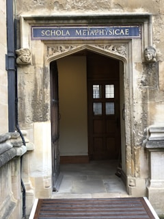 An ornate stone doorway with a sign reading 'SCHOLA METAPHYSICAE' in uppercase letters above. The doorway is framed by carved stone faces on each side. The interior visible through the doorway features wooden panels and a window with small panes. The stonework is weathered, giving a historical appearance.