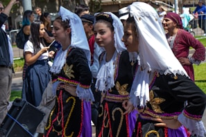 Several women are dressed in traditional, ornate costumes with intricate embroidery and lace. They are part of a gathering, possibly a cultural festival or parade, with other people visible in the background, some of whom are also in costume.