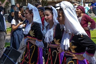 Several women are dressed in traditional, ornate costumes with intricate embroidery and lace. They are part of a gathering, possibly a cultural festival or parade, with other people visible in the background, some of whom are also in costume.