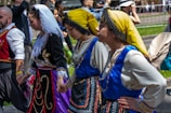 A group of people in traditional folk costumes are holding hands in a line during an outdoor event. The costumes are elaborate, with one featuring a yellow headscarf and blue embellishments, another adorned with gold embroidery and a white headscarf. The scene is vibrant and lively, with onlookers in the background, some capturing the moment.