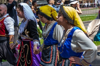 A group of people in traditional folk costumes are holding hands in a line during an outdoor event. The costumes are elaborate, with one featuring a yellow headscarf and blue embellishments, another adorned with gold embroidery and a white headscarf. The scene is vibrant and lively, with onlookers in the background, some capturing the moment.