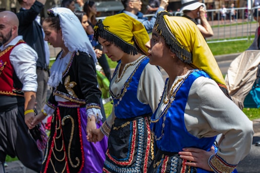 A group of people in traditional folk costumes are holding hands in a line during an outdoor event. The costumes are elaborate, with one featuring a yellow headscarf and blue embellishments, another adorned with gold embroidery and a white headscarf. The scene is vibrant and lively, with onlookers in the background, some capturing the moment.