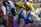 A group of people in traditional folk costumes are holding hands in a line during an outdoor event. The costumes are elaborate, with one featuring a yellow headscarf and blue embellishments, another adorned with gold embroidery and a white headscarf. The scene is vibrant and lively, with onlookers in the background, some capturing the moment.