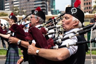 A group of musicians are playing bagpipes outdoors. They are dressed in traditional Scottish attire, including kilts and hats adorned with feathers and badges. The background features urban buildings and a clear sky, suggesting a city setting.