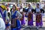 A group of young women is dressed in traditional colorful costumes, participating in what seems to be a cultural parade or festival. They are wearing elaborately designed dresses with intricate patterns and headscarves. The background shows more participants and spectators alongside a street.