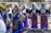 A group of young women is dressed in traditional colorful costumes, participating in what seems to be a cultural parade or festival. They are wearing elaborately designed dresses with intricate patterns and headscarves. The background shows more participants and spectators alongside a street.
