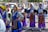 A group of young women is dressed in traditional colorful costumes, participating in what seems to be a cultural parade or festival. They are wearing elaborately designed dresses with intricate patterns and headscarves. The background shows more participants and spectators alongside a street.