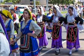 A group of young women is dressed in traditional colorful costumes, participating in what seems to be a cultural parade or festival. They are wearing elaborately designed dresses with intricate patterns and headscarves. The background shows more participants and spectators alongside a street.