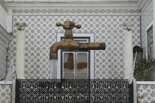 A large brass faucet appears to float in midair against a decorative patterned wall. It is attached to a structure framed by two columns and ornate ironwork. Water is visibly flowing from the faucet, adding a surreal element to the scene.