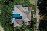 An aerial view of a serene tropical villa surrounded by lush greenery. The villa features traditional thatched roofs with red accents and a rectangular swimming pool. A path leads through well-manicured gardens filled with palm trees and vibrant plants. There are umbrellas and seating areas around the pool area, providing a tranquil, vacation-like atmosphere.