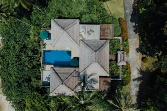 An aerial view of a serene tropical villa surrounded by lush greenery. The villa features traditional thatched roofs with red accents and a rectangular swimming pool. A path leads through well-manicured gardens filled with palm trees and vibrant plants. There are umbrellas and seating areas around the pool area, providing a tranquil, vacation-like atmosphere.