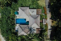 An aerial view of a serene tropical villa surrounded by lush greenery. The villa features traditional thatched roofs with red accents and a rectangular swimming pool. A path leads through well-manicured gardens filled with palm trees and vibrant plants. There are umbrellas and seating areas around the pool area, providing a tranquil, vacation-like atmosphere.