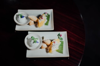 Two rectangular plates each hold a stylish dessert arrangement, featuring a scoop of white ice cream garnished with a green mint leaf and a tiny purple flower, alongside pastries and a piece of fruit adorned with seeds. The plates are presented on a dark wooden surface with a dimly lit background, adding an elegant touch to the scene.
