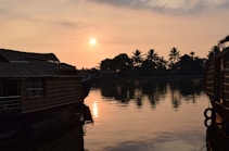 The image captures a serene sunset over a calm body of water, with silhouettes of houseboats in the foreground. The sun is setting in the clear sky, casting a warm golden glow over the scene. Palm trees and other foliage are visible along the horizon, reflected in the tranquil water surface.