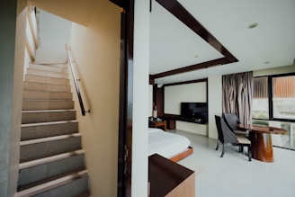 A modern interior with a staircase on the left leading upwards, adjacent to a bedroom area. The bedroom features a neatly made bed, a flat-screen television mounted on the wall, and a workspace with a wooden desk and a chair. To the right, large windows with drapes allow natural light to fill the room.