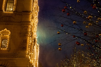 A beautifully decorated building facade is illuminated with golden lights. To the right, a tree is adorned with red and gold ornaments, with its branches outlined by small white lights. The background features a deep blue night sky, enhancing the festive atmosphere.